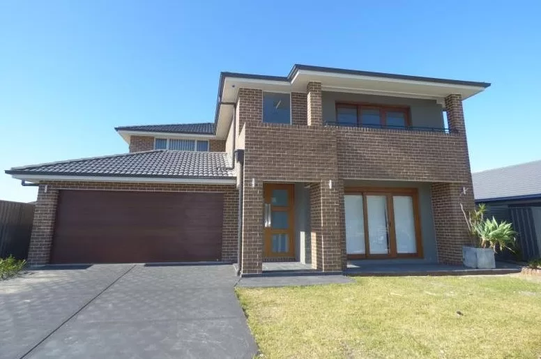 The image shows a modern two-story house with a brick exterior. The house features a large double garage with wooden doors on the left side and a main entrance with a wooden door and glass panels in the center. There is a balcony on the second floor above the entrance, supported by brick columns. The house has a tiled roof and large windows, including sliding glass doors on the ground floor. The front yard has a small lawn area and a concrete driveway leading to the garage. There are also some plants in a planter on the right side near the entrance. The sky is clear and blue, indicating a sunny day. This image is interesting as it showcases a contemporary residential property, which could be of interest to potential homebuyers, real estate agents, or architects looking for design inspiration.